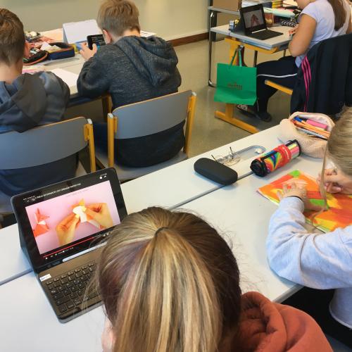 Schülerinnen und Schüler malen ihr Papier in Herbstfarben an, eine Schülerin sieht sich am iPad eine Faltanleitung für ein Origami-Tier an